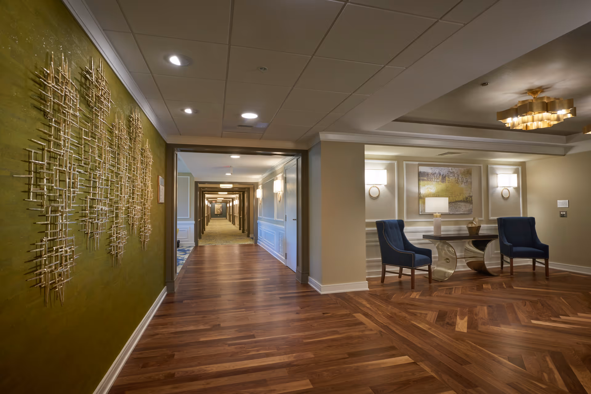 Interior lobby corridor with hardwood floors, decorative wall art on the left, and a seating area with two blue chairs and a console table on the right.