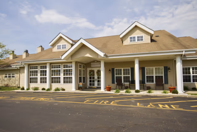 Exterior front view of Whitlock Place, a single-story building with beige siding, white columns, and multiple windows. The entrance has a covered porch with two chairs and potted plants. The parking area in front has yellow painted markings indicating a fire lane.
