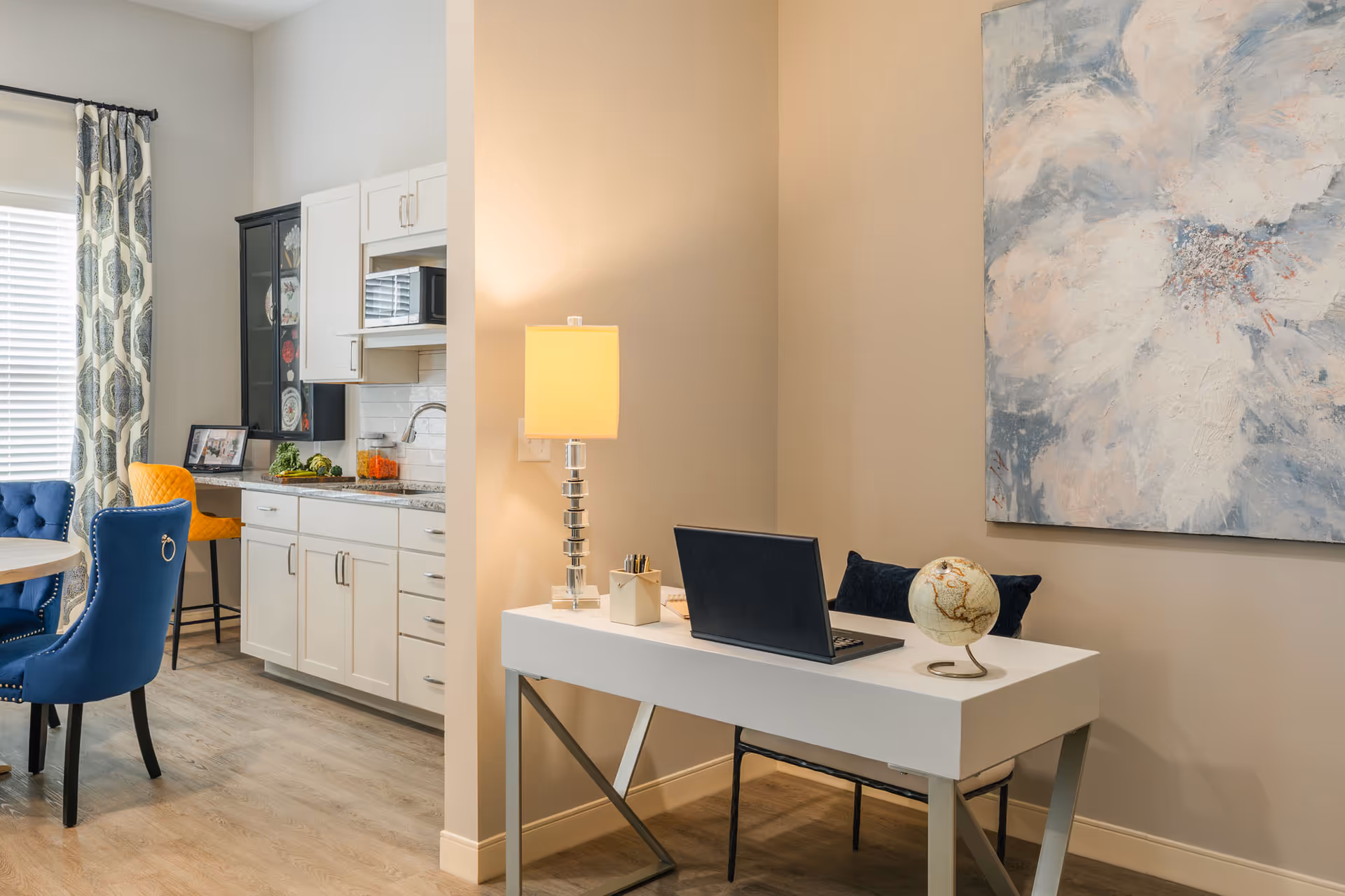 A modern interior space featuring a small white desk with a laptop, a globe, and a lamp. Behind the desk is a large abstract painting on the wall. To the left, there is a kitchenette with white cabinets, a countertop, and a microwave. A dining area with blue and yellow upholstered chairs and a round table is partially visible near a window with patterned curtains.