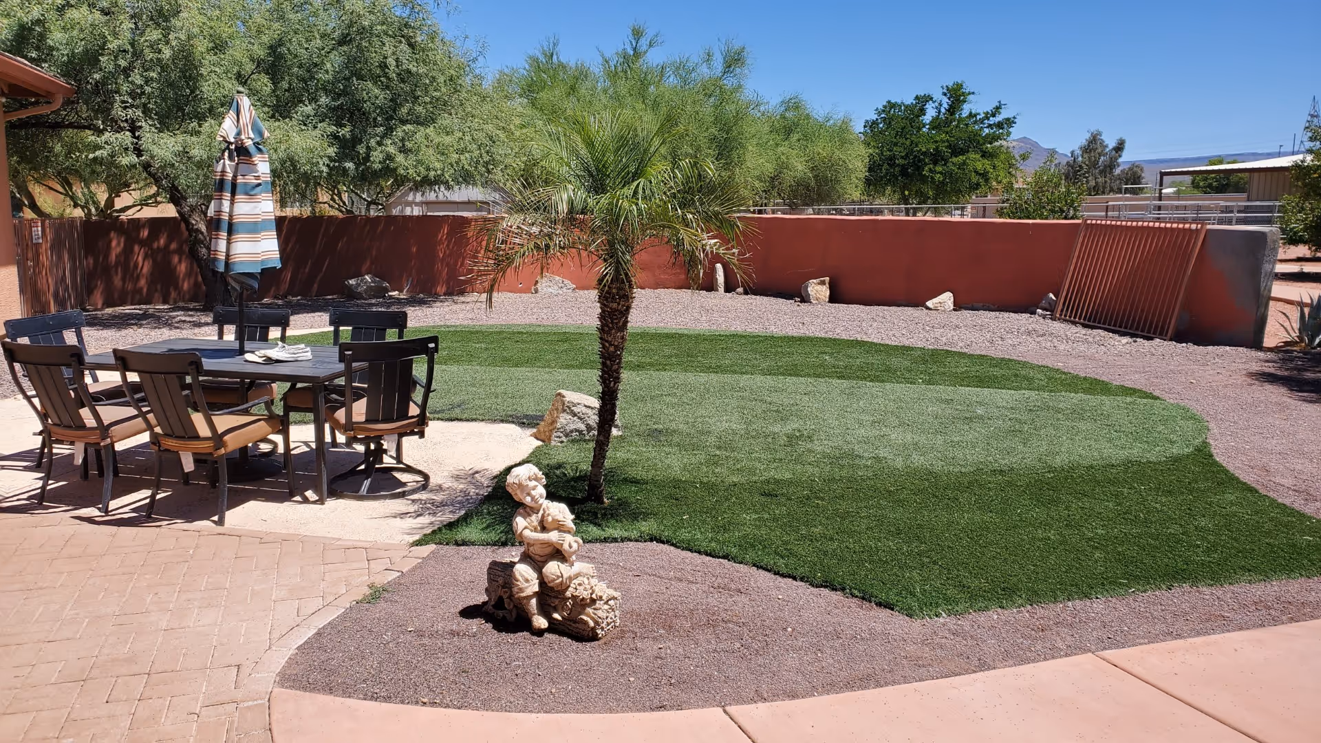 Outdoor patio area with a table and six chairs, a closed striped umbrella, a small palm tree, and a statue of a child sitting on a log. The ground is a mix of paved walkway, gravel, and patches of artificial grass. Trees and a red wall surround the area under a clear blue sky.