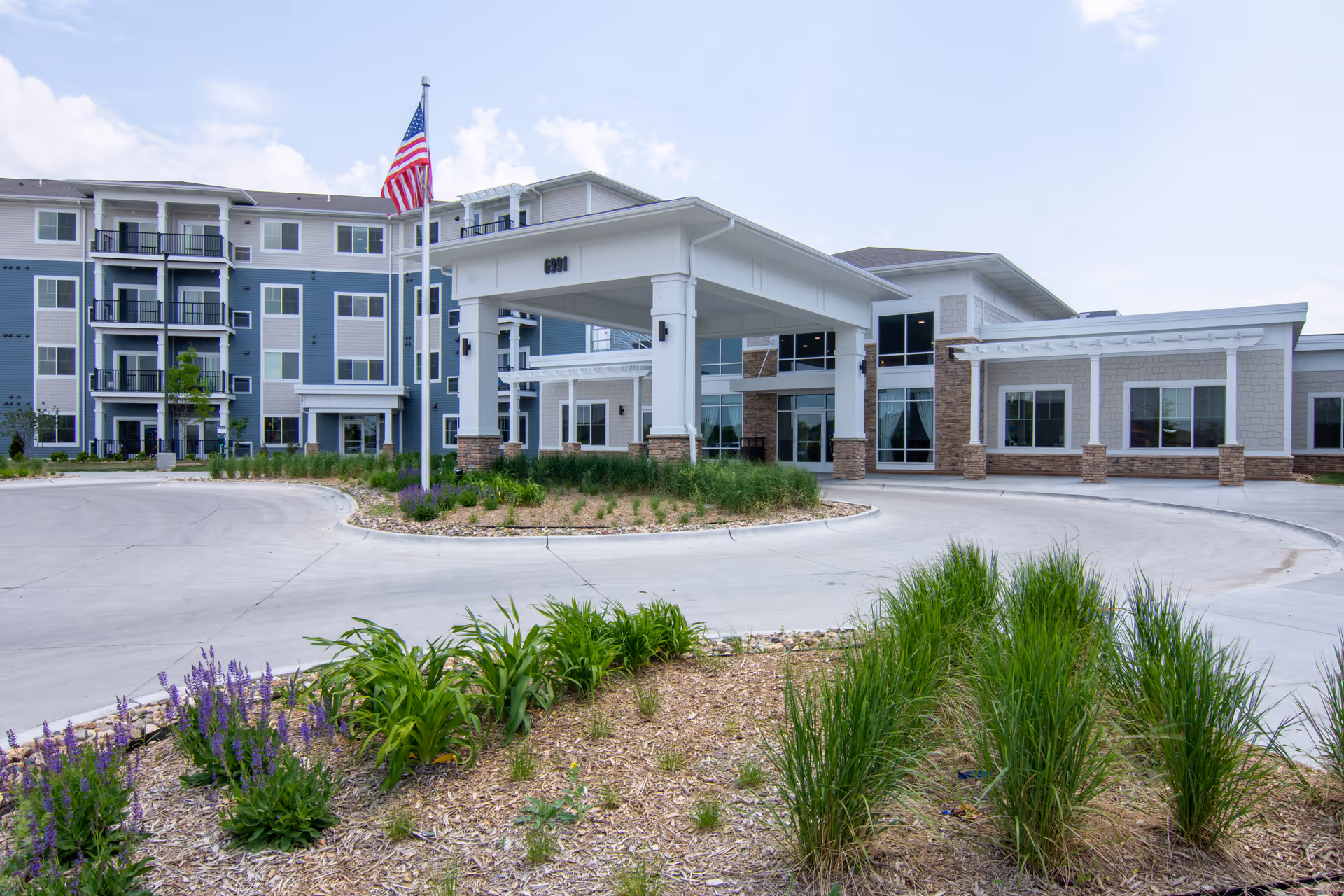 Front exterior view of The Brant at Gretna Senior Living facility showing a modern multi-story building with balconies, an American flag on a flagpole, a covered entrance with pillars, and landscaped greenery in the foreground under a partly cloudy sky.