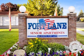 A brick sign for Fort Lane Senior Apartments with an American flag graphic, located at 300 North Fort Lane, surrounded by colorful flowers and greenery with a clear sky in the background.