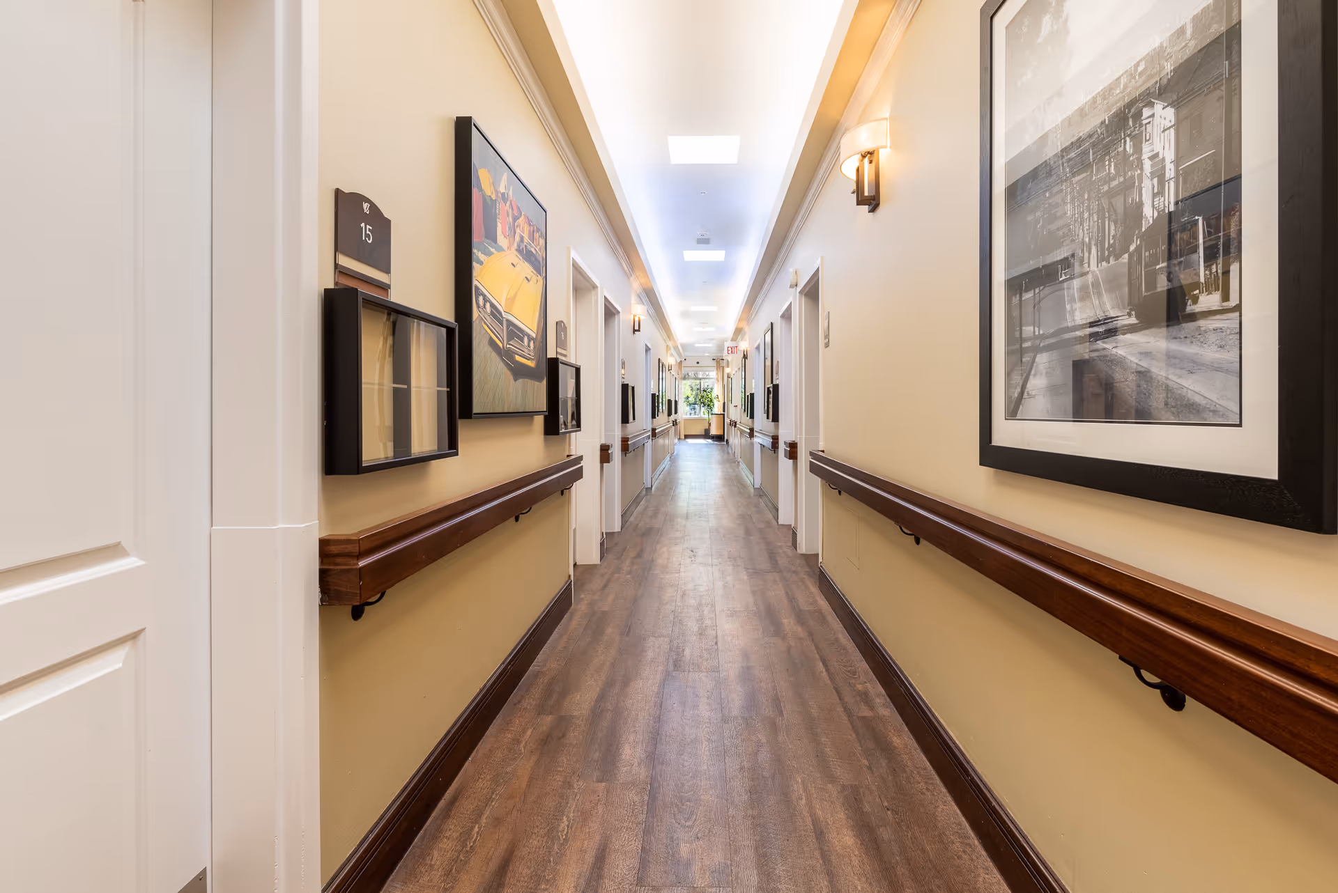 Well-lit interior corridor with wood handrails, framed artwork, and doors lining both sides.