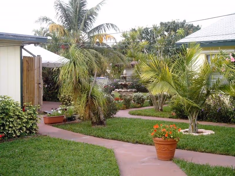 A garden area with a winding paved walkway surrounded by green grass, palm trees, and various plants. There is a terracotta pot with orange flowers in the foreground and a multi-tiered water fountain in the background. Buildings with light-colored walls and a wooden gate are visible on either side.