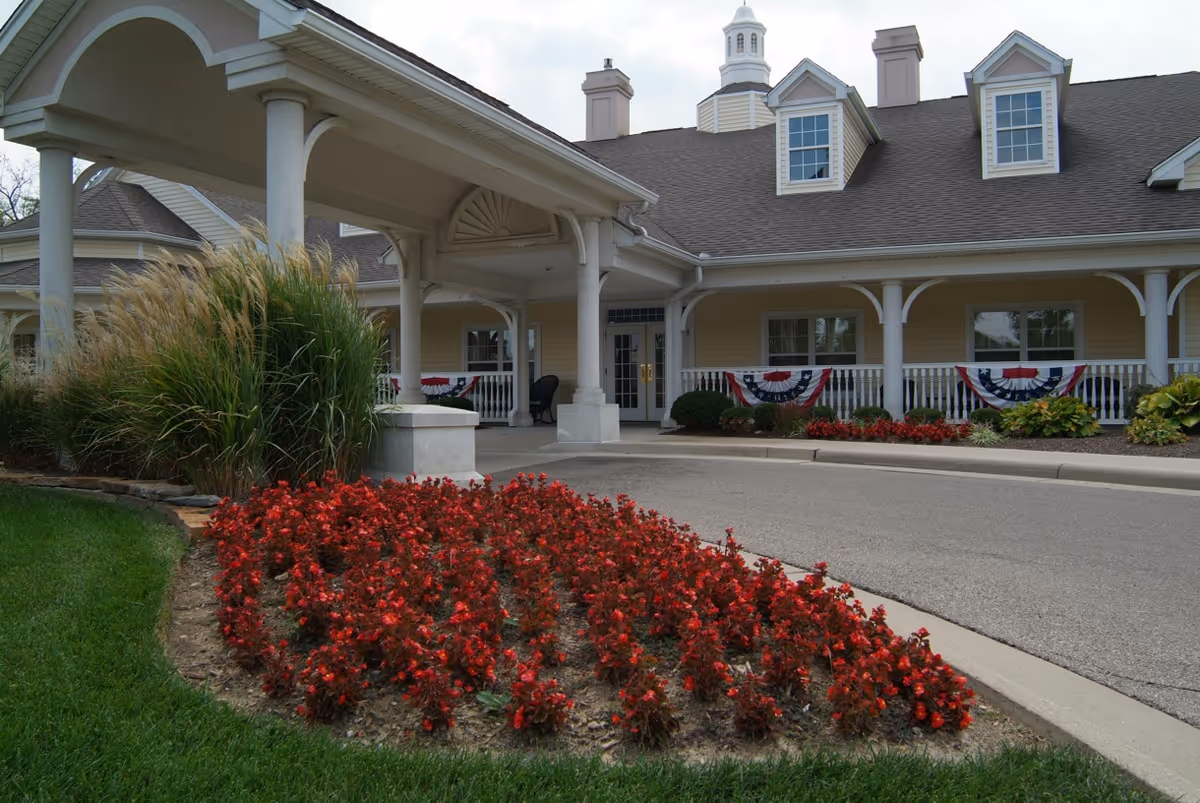 Front exterior view of a senior living facility with a covered entrance, manicured flower beds with red flowers, ornamental grasses, and patriotic bunting decorations on the porch railing.
