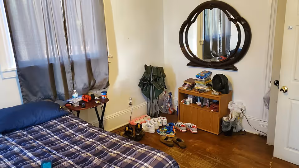 Bedroom with a plaid-covered bed, small side table, dresser under a wall mirror and several pairs of shoes on the floor.