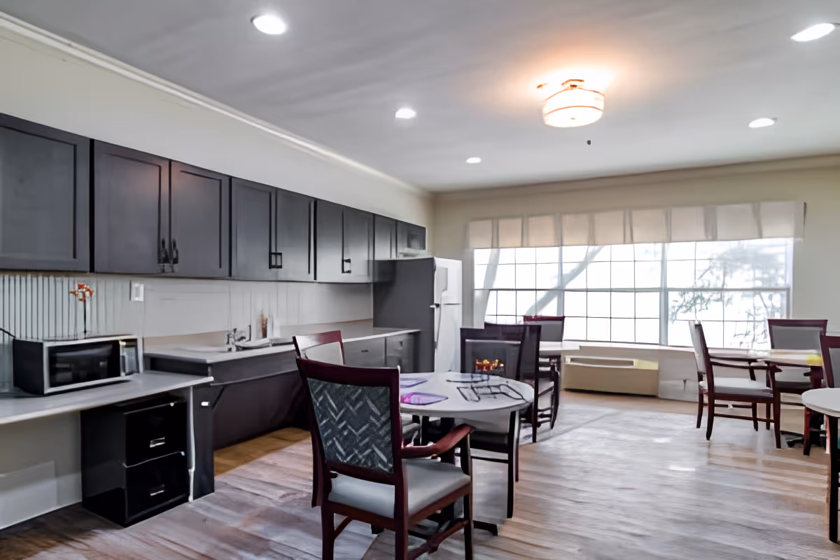 Bright communal dining room with a kitchenette, round tables and chairs, and large windows.