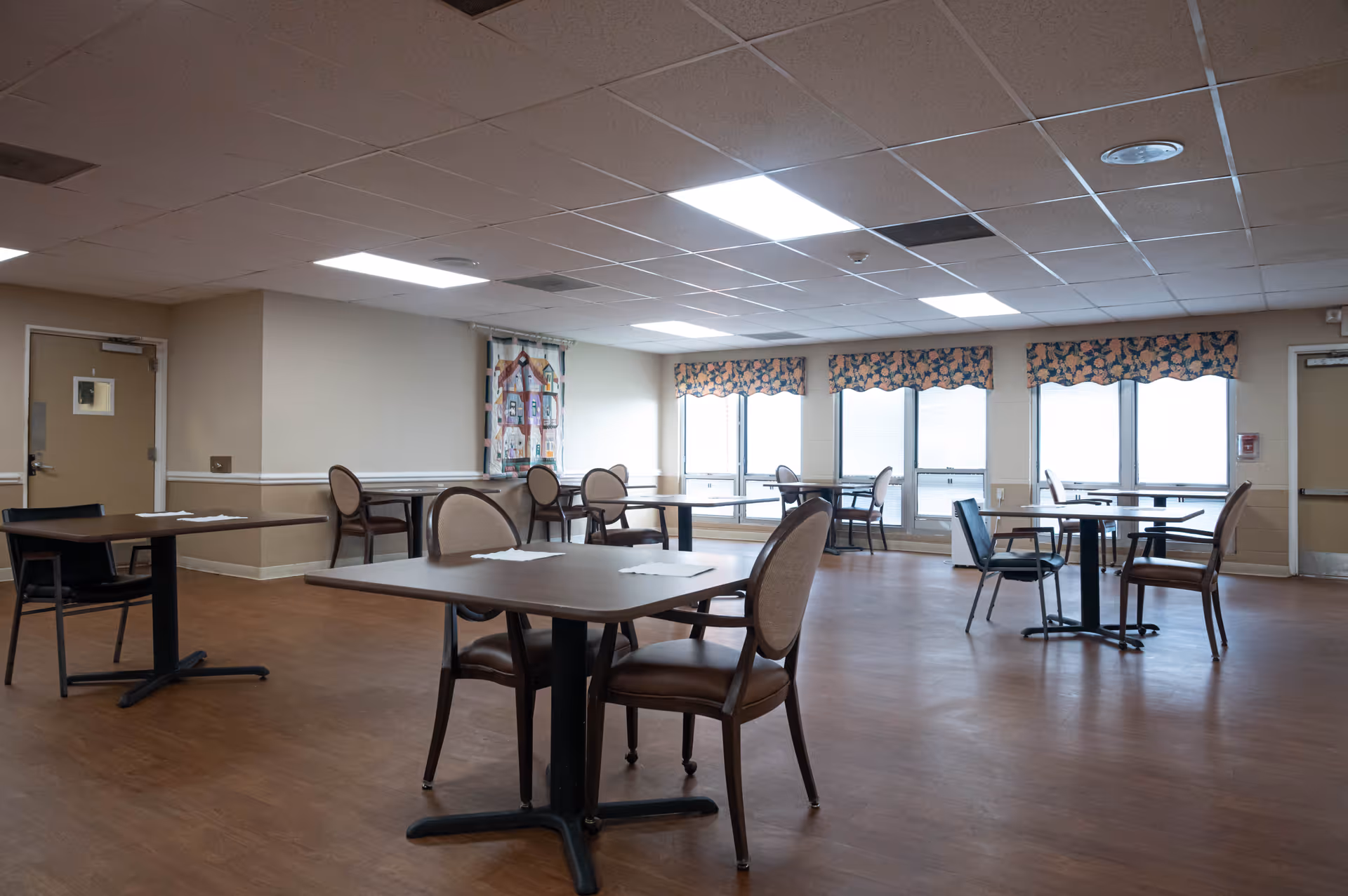 Spacious communal dining room with several tables and chairs and large windows along one wall.