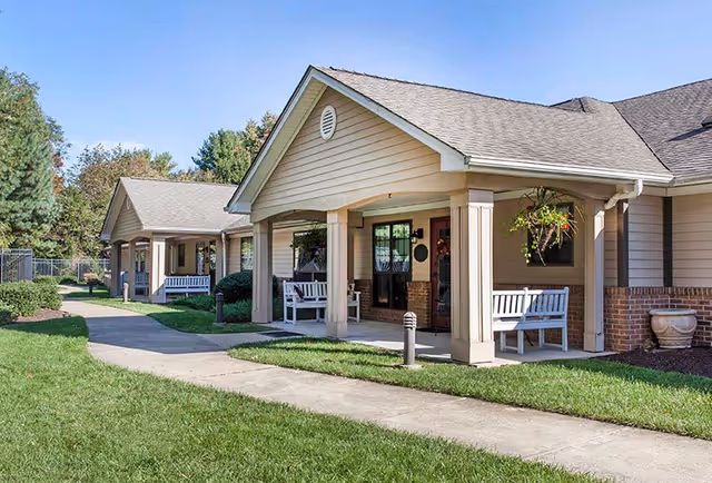 Exterior view of a senior living facility with beige siding and brick accents, featuring covered porches with white benches and hanging plants, a paved walkway, green grass, and trees in the background under a clear blue sky.