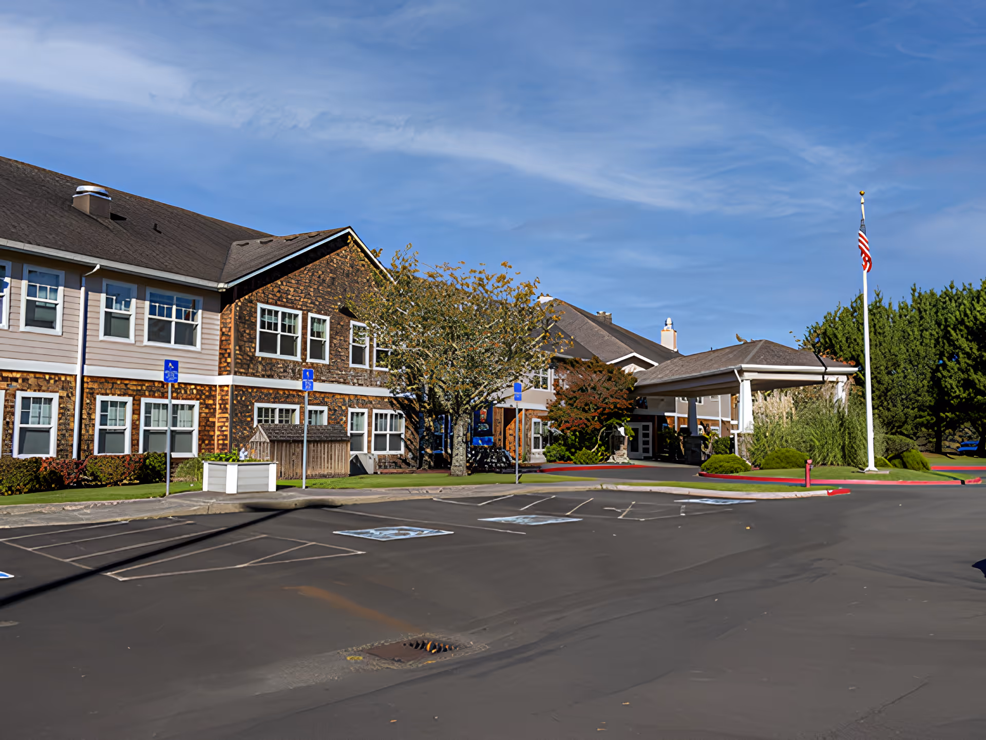 Exterior view of Avamere at Seaside senior living facility showing a two-story building with a covered entrance, several windows, trees, and an American flag on a flagpole. The parking lot in front has marked handicap parking spaces.