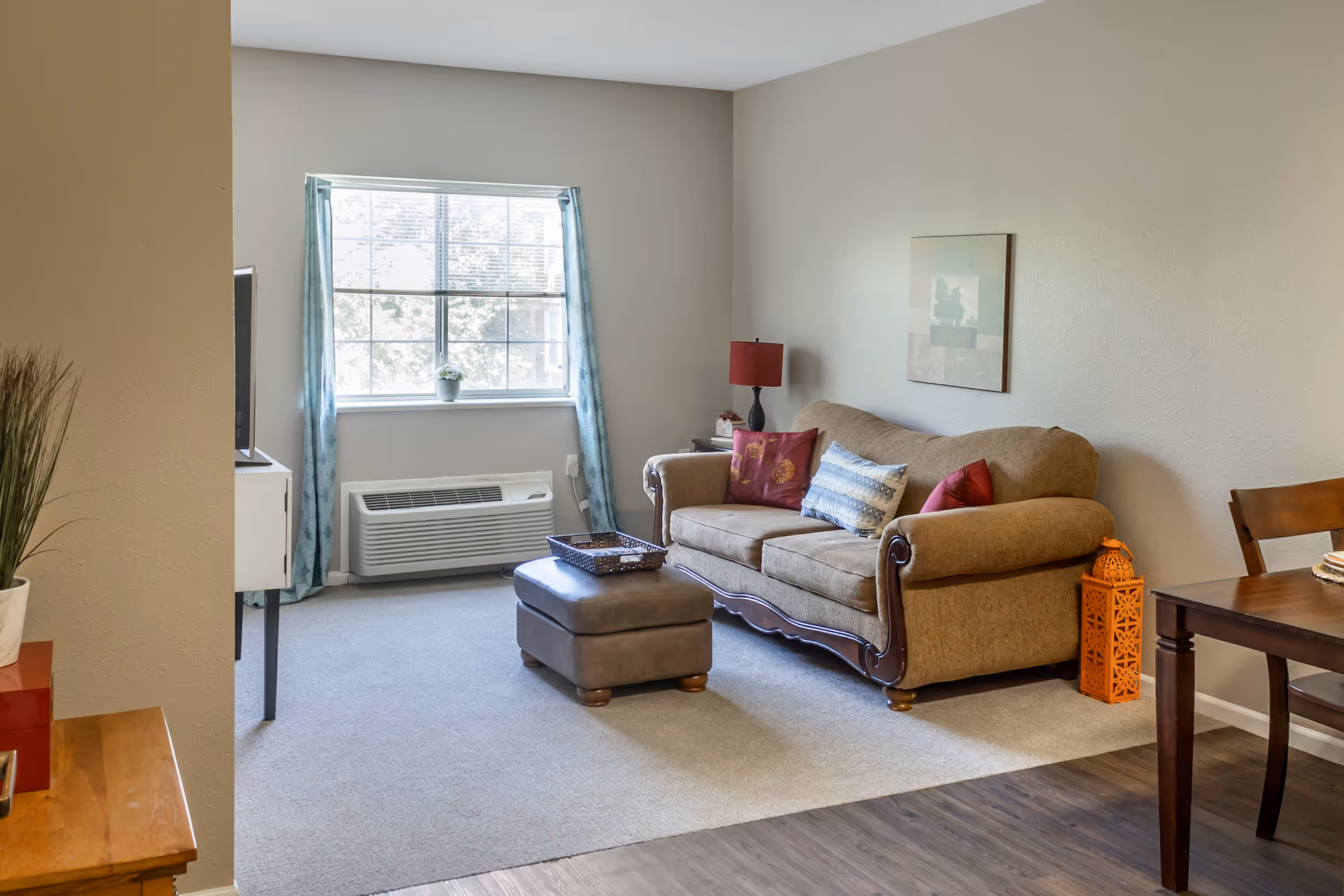 Cozy living room with a brown sofa, ottoman, TV stand, window with blue curtains, and a small dining table.