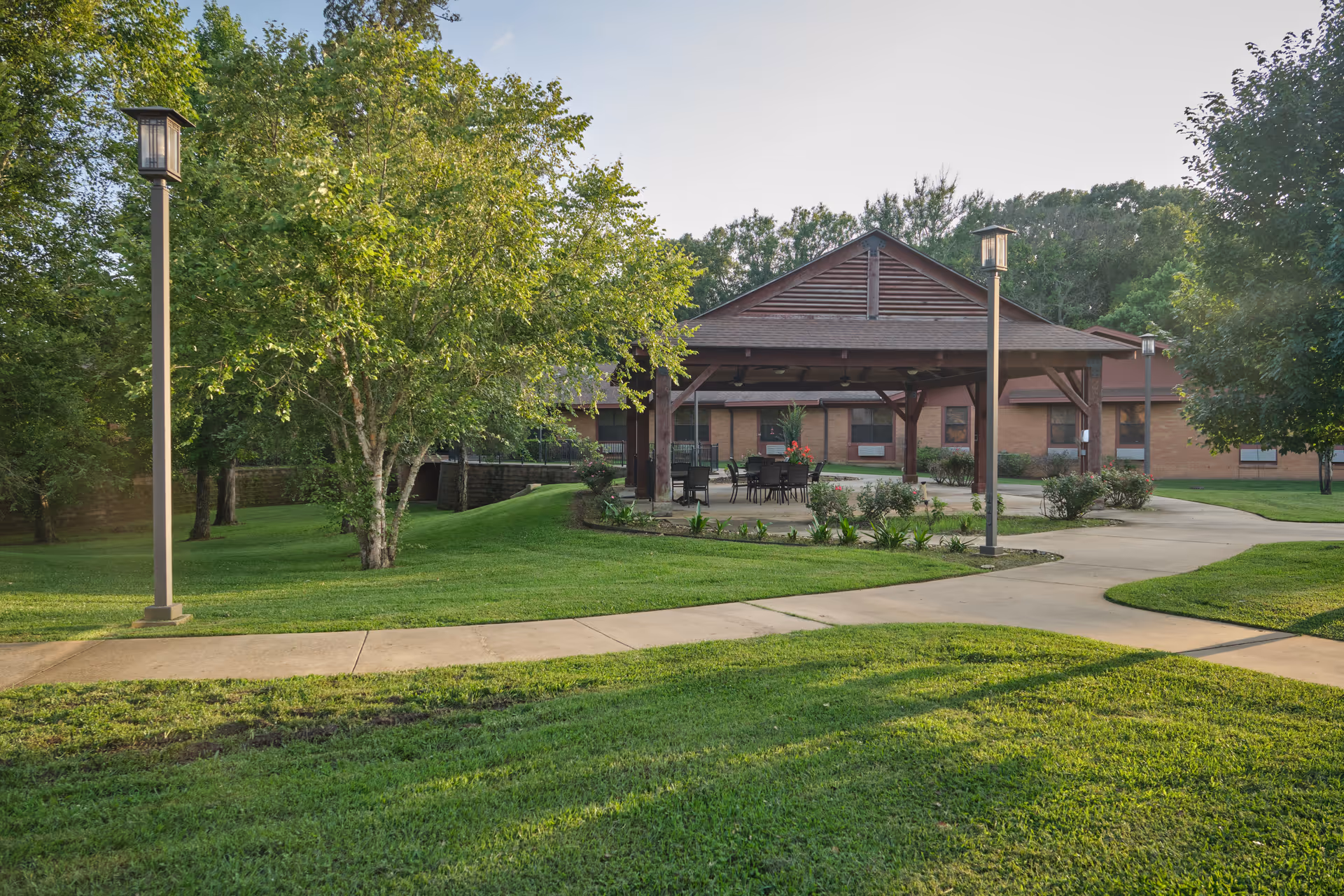 Covered outdoor pavilion with tables and chairs in a grassy courtyard in front of a brick senior living building.