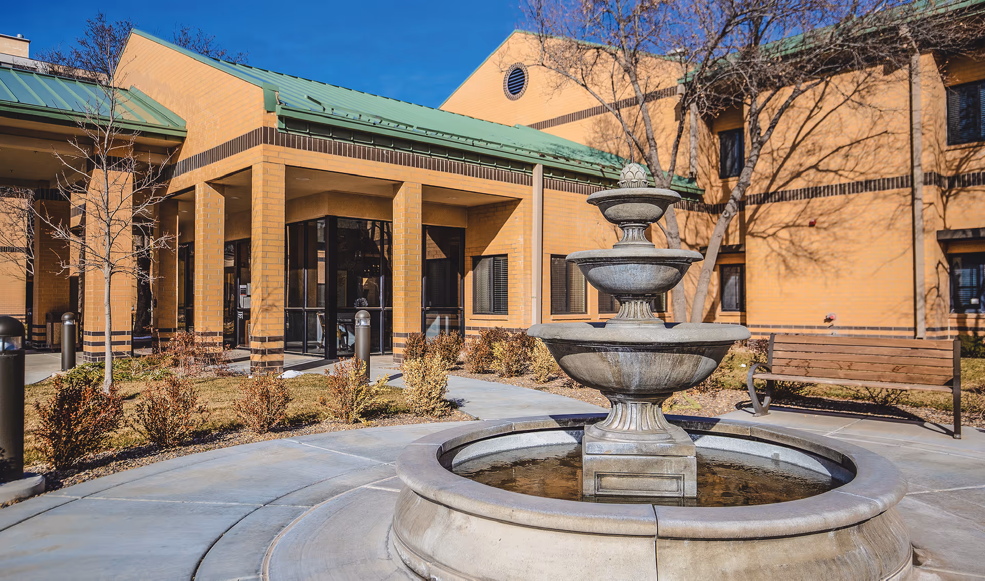 Courtyard front entrance of Park Central Community showing a multi-tiered stone fountain, bench, and the building facade.