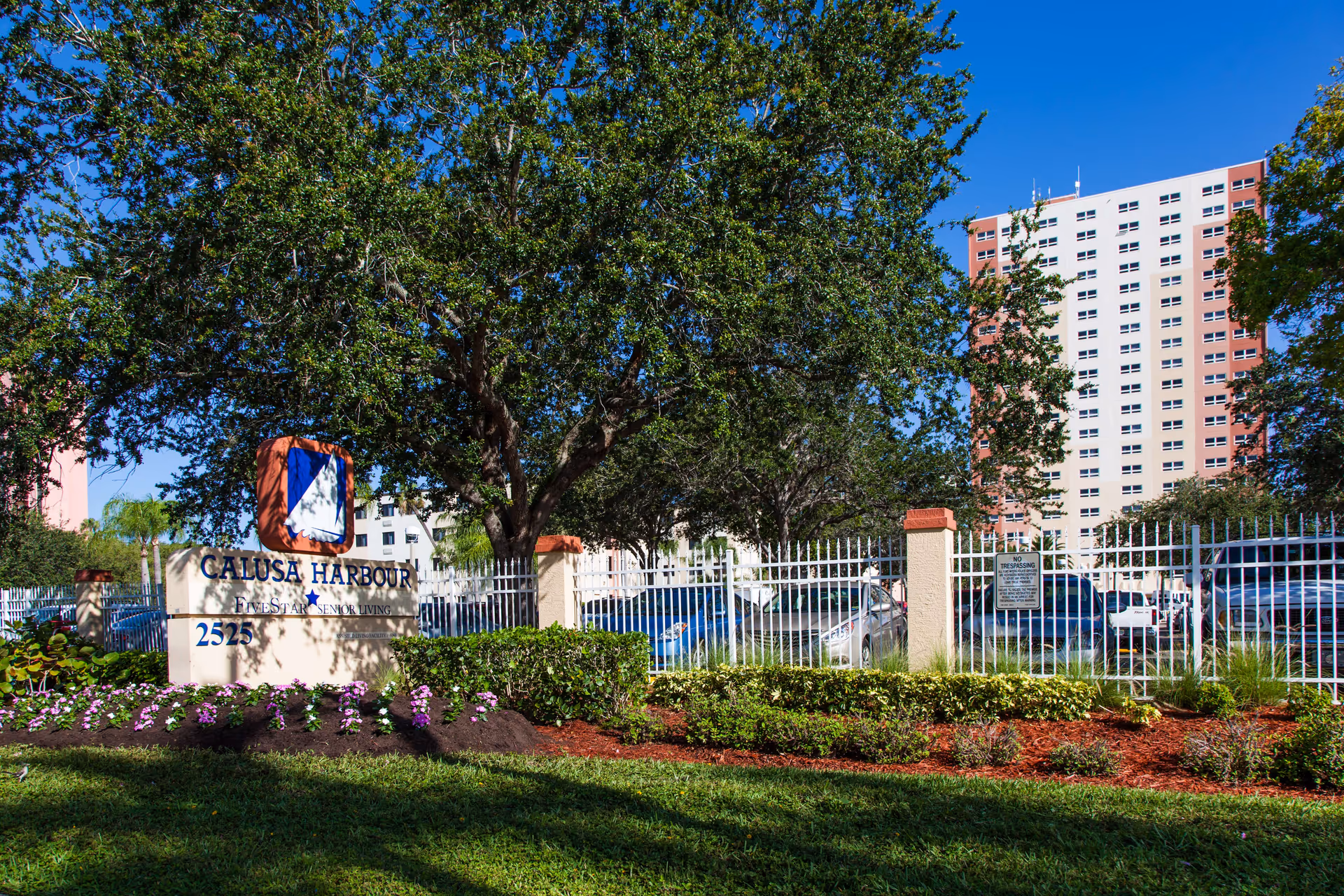Exterior view of Calusa Harbour senior living facility with a sign displaying the name and address, surrounded by trees, flowers, and a white fence. A tall building and parked cars are visible in the background under a clear blue sky.