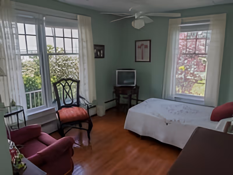 A cozy bedroom with light green walls, a bed covered with a white bedspread, a small vintage TV on a wooden table, a wooden chair with a red cushion, a red armchair, and large windows with sheer white curtains letting in natural light.