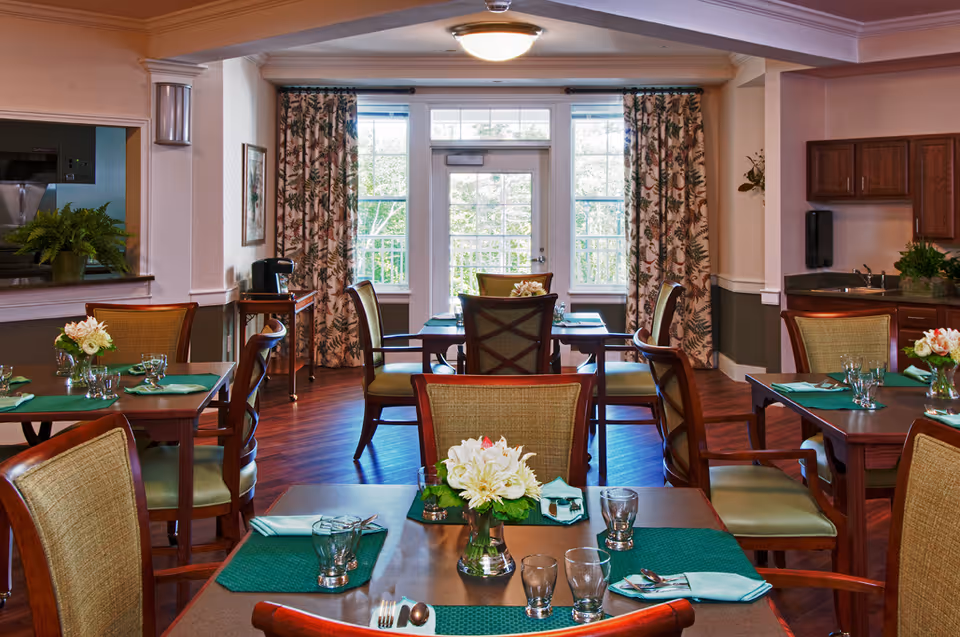 A bright dining room with multiple set tables and wooden chairs, floral centerpieces, and a door with windows framed by patterned curtains.