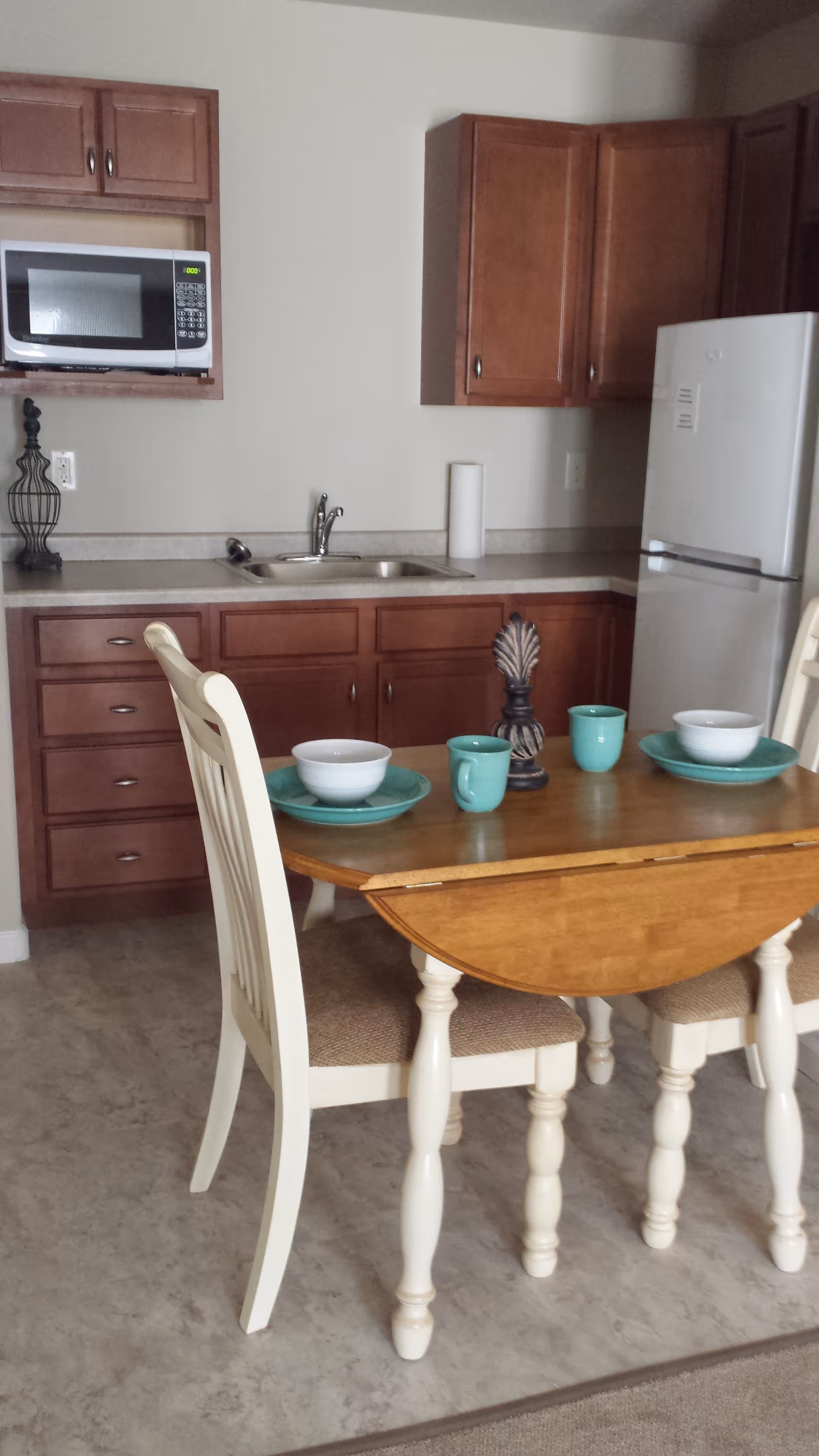 A small kitchen area with wooden cabinets, a white microwave mounted above the counter, a stainless steel sink, and a white refrigerator. In front of the kitchen is a wooden dining table with two white chairs. The table is set with two turquoise plates, two white bowls, and two turquoise mugs, along with a decorative centerpiece.
