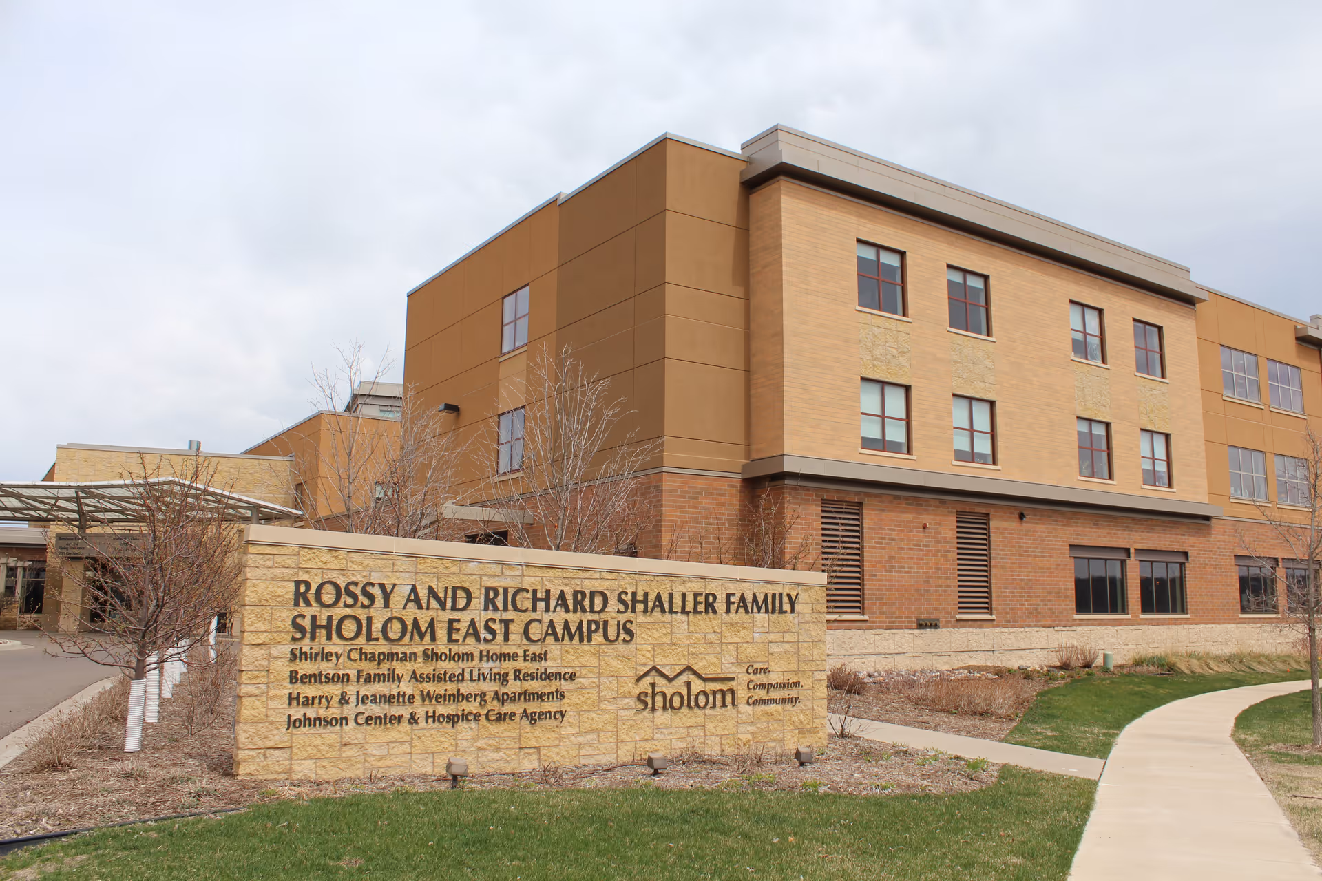 Exterior view of the Rossy and Richard Shaller Family Sholom East Campus building with a stone sign in front listing various facilities including Shirley Chapman Sholom Home East, Bentson Family Assisted Living Residence, Harry & Jeanette Weinberg Apartments, and Johnson Center & Hospice Care Agency. The building is three stories tall with multiple windows and a pathway leading to the entrance.