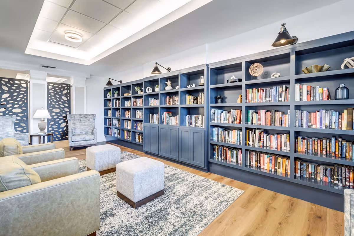 A bright and modern library or reading room with a large blue built-in bookshelf filled with books and decorative items. The room features comfortable seating including armchairs and ottomans on a patterned rug, with soft lighting and decorative panels in the background.
