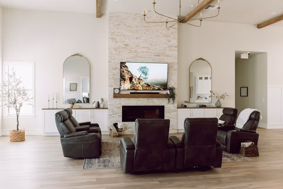 A spacious living room with four black leather recliners arranged around a stone fireplace with a mounted flat-screen TV displaying a nature scene. The room features light wood flooring, two large decorative mirrors above white cabinets on either side of the fireplace, a potted plant near a window, and a modern chandelier hanging from the ceiling with exposed wooden beams.