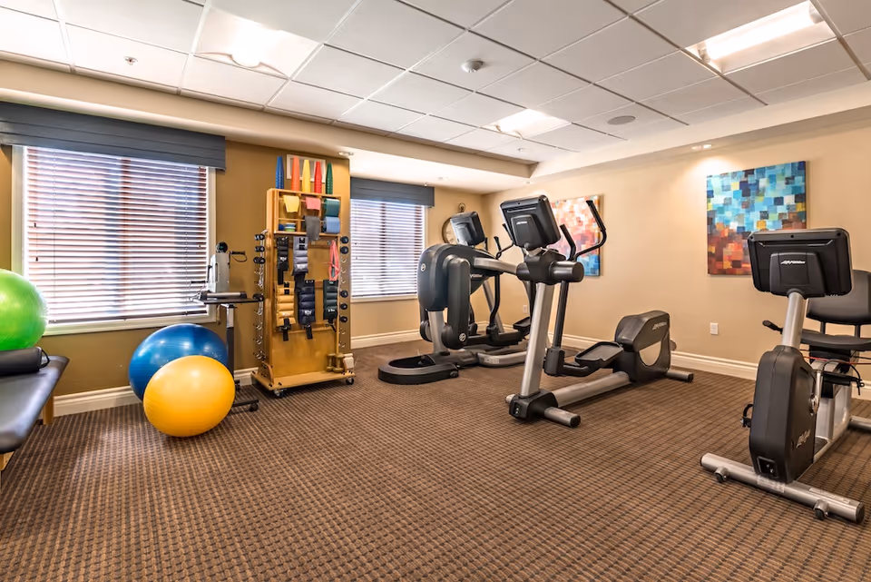 A senior living facility fitness room with exercise equipment including stationary bikes, elliptical machines, exercise balls, and a rack holding various fitness accessories. The room has carpeted flooring, two windows with blinds, and colorful abstract paintings on the walls.