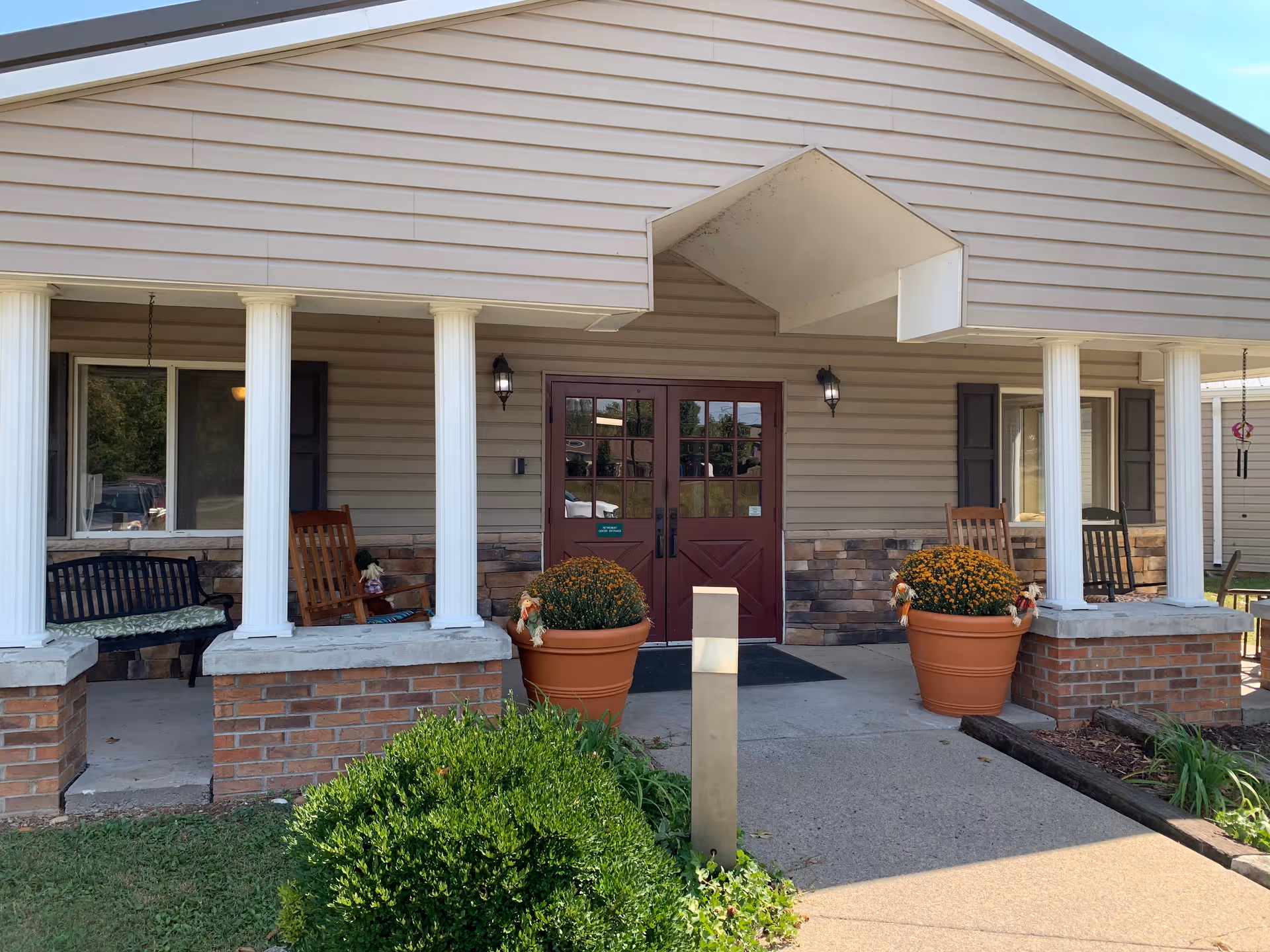Front entrance of a single-story retirement community building with a covered porch, white columns, double red doors and large potted flowers.
