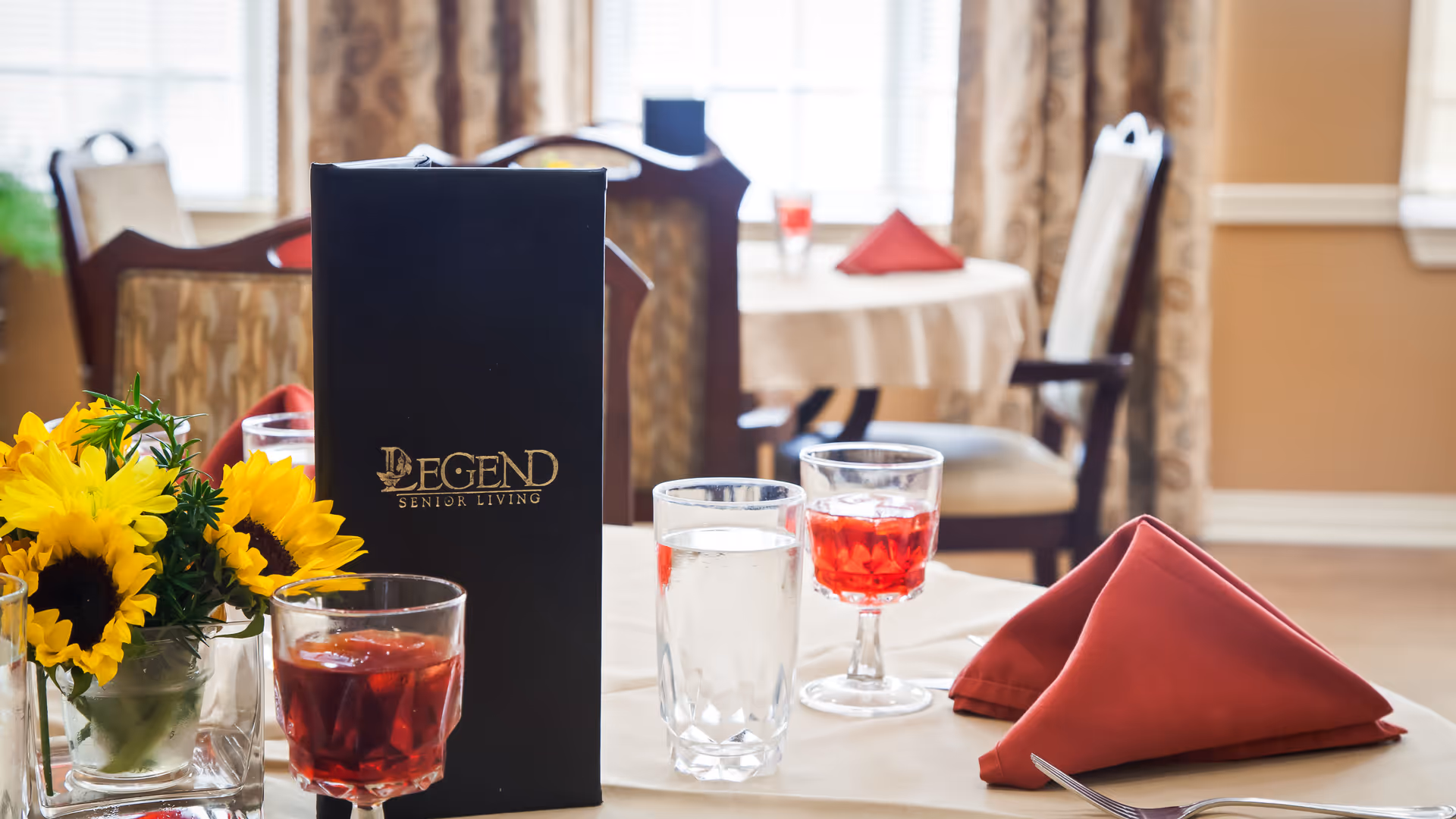 A dining table set with a black menu labeled 'Legend Senior Living,' two glasses filled with water and red beverage, a folded red napkin, and a small vase of yellow sunflowers. In the background, there are more tables and chairs with similar settings in a warmly lit dining room.