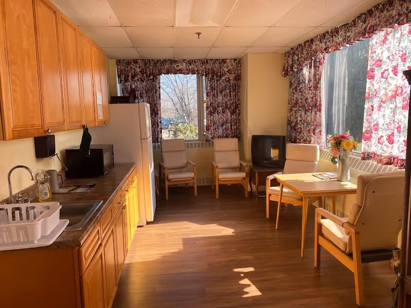 A bright room with wooden cabinets and a countertop on the left side, including a sink, microwave, and refrigerator. On the right side, there is a small table with a vase of flowers and four cushioned chairs. The room has large windows with floral curtains letting in natural light, and a small TV is placed on a stand near the back wall.