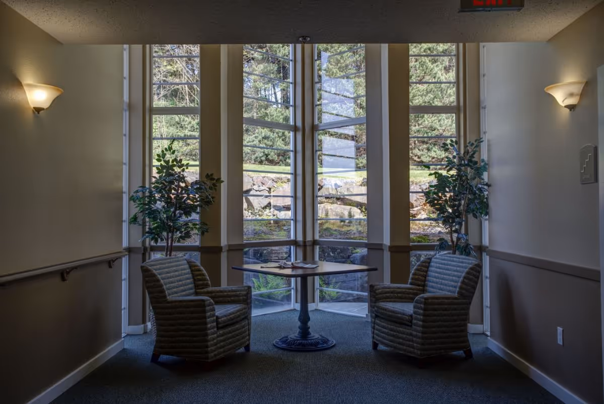 A cozy seating area in a hallway with two striped armchairs facing each other across a small round table. The space is illuminated by wall sconces and natural light coming through tall windows that show a view of greenery and rocks outside. There are two potted plants on either side of the windows.