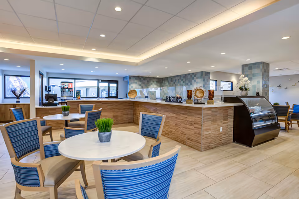 Bright and modern dining area with round white tables and blue-striped chairs. A counter labeled 'Bistro' with decorative items and a refrigerated display case is visible. The space has tiled flooring, recessed lighting, and large windows providing natural light.
