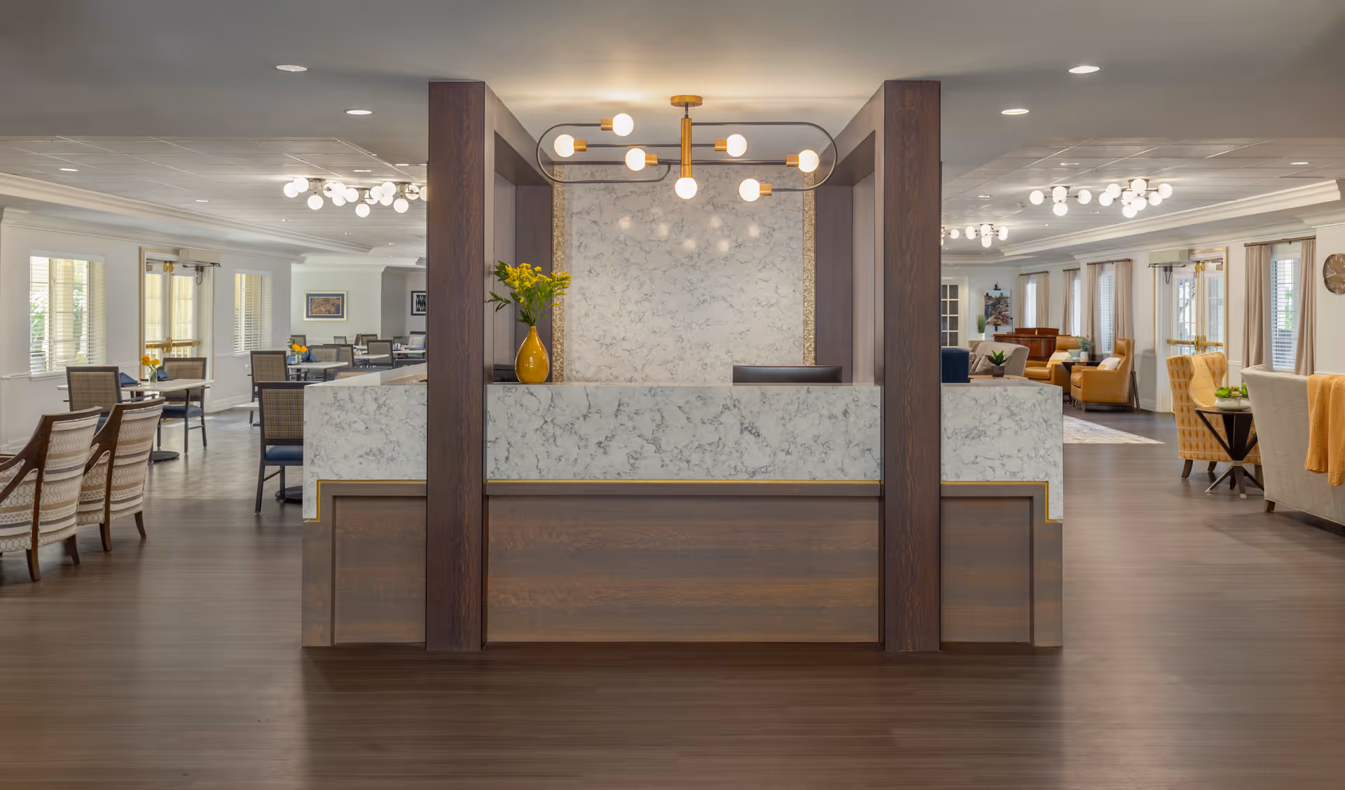 A spacious and well-lit reception area with a modern marble and wood front desk. Behind the desk is a decorative wall with a contemporary light fixture. The surrounding area features multiple seating arrangements with chairs and tables, large windows with blinds, and additional lighting fixtures on the ceiling.