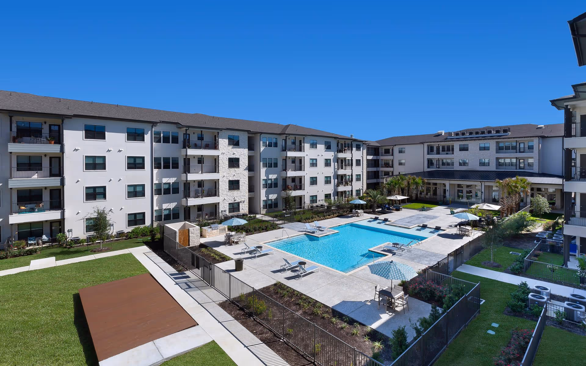 Outdoor courtyard area of a senior living facility with a large swimming pool surrounded by lounge chairs and umbrellas. The pool is enclosed by a black metal fence, and there are multiple four-story residential buildings with balconies surrounding the courtyard. The sky is clear and blue.