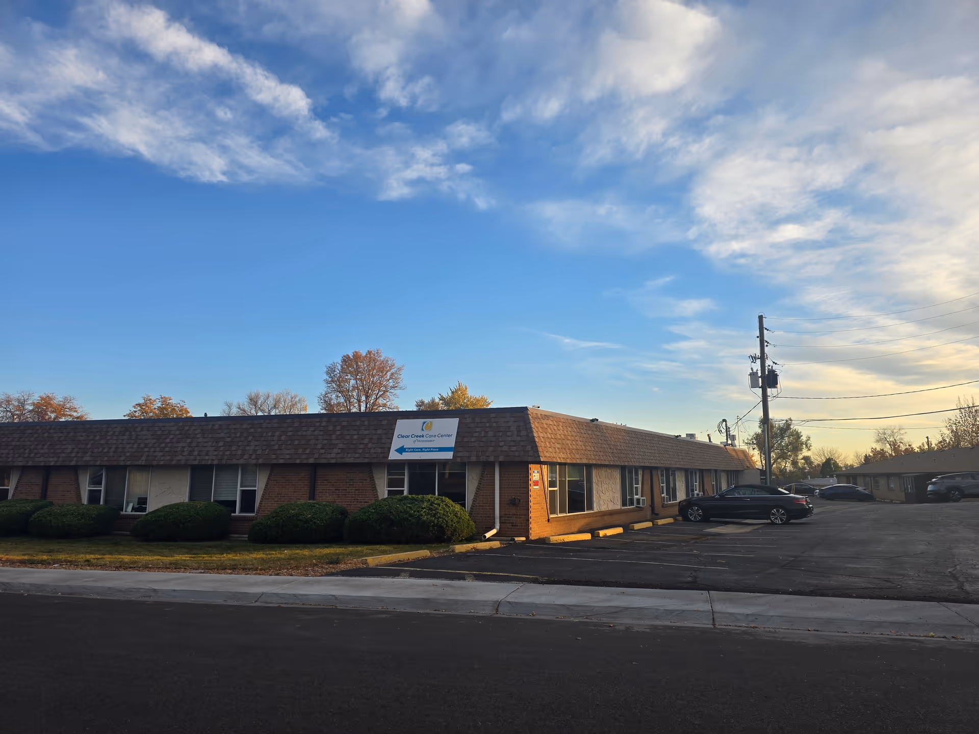 Exterior view of Clear Creek Care Center, a single-story brick building with a shingled roof, surrounded by bushes and a parking lot with several cars. The sky is partly cloudy with a blue and light yellow hue.
