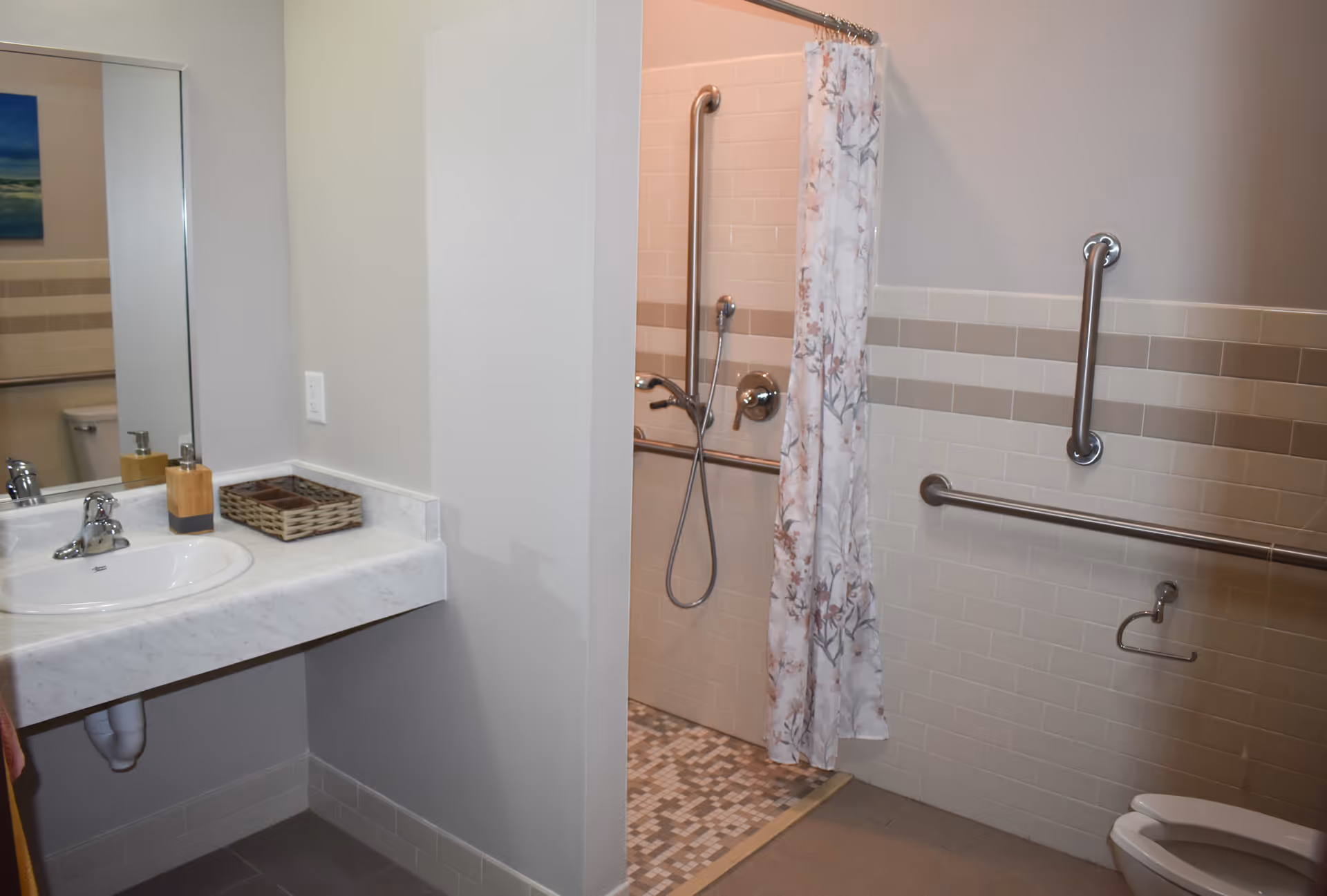 A bathroom with a sink and mirror on the left side, a walk-in shower with a floral shower curtain in the center, and a toilet on the right. The walls have beige and white tiles with grab bars installed for accessibility.