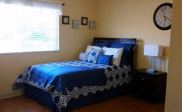 A neatly made single bed with blue patterned bedding and pillows next to a nightstand with a lamp, a wall clock, and a window in a small bedroom.