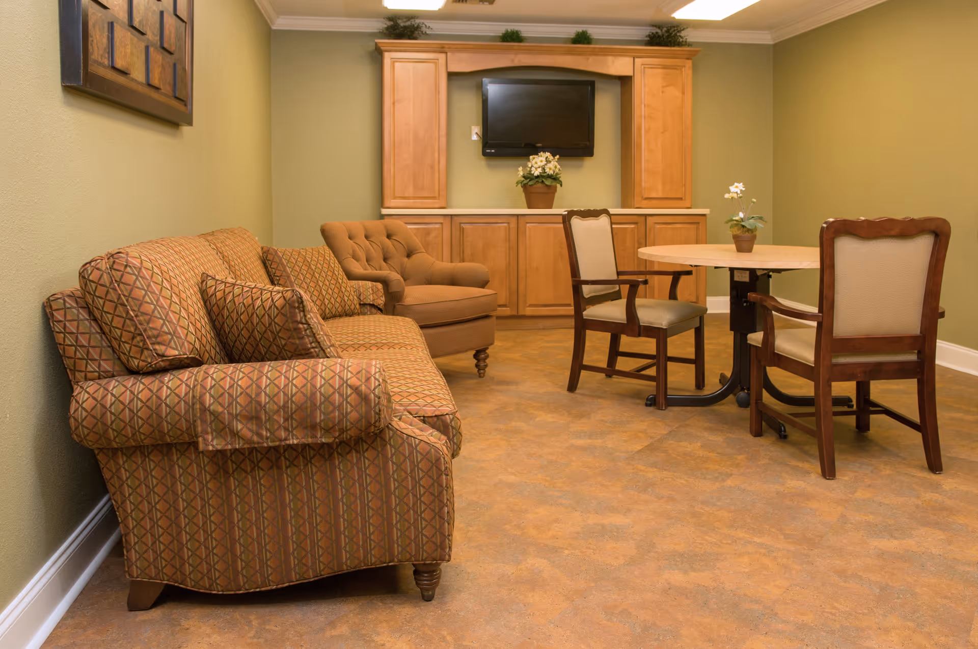 A cozy living room area with a patterned sofa, a tufted armchair, and a round wooden table with two cushioned chairs. The walls are painted light green, and there is a wooden cabinet with a flat-screen TV mounted above it. Small potted plants are placed on the cabinet and table.