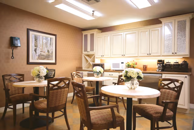 Small communal dining area with round tables and chairs, floral centerpieces, and a kitchenette with cabinets and appliances in the background.