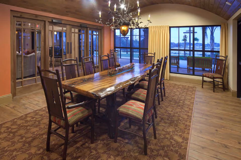 A dining room with a long wooden table surrounded by ten chairs with striped cushions. The room has wooden floors, a patterned rug under the table, and a large chandelier hanging above. Large windows with yellow curtains offer a view of an outdoor patio and trees beyond.