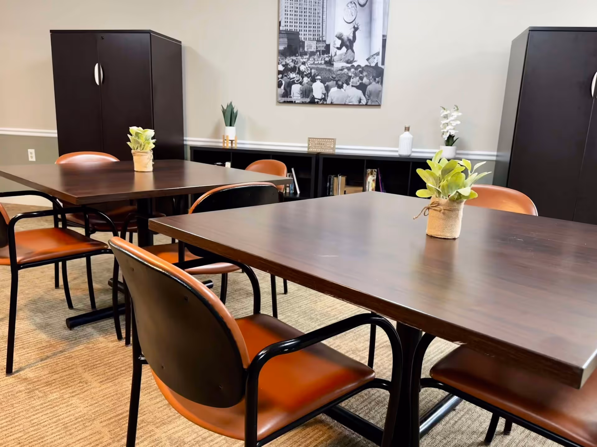 A room with two dark wooden tables each decorated with a small potted plant. Surrounding the tables are brown chairs with black metal frames. In the background, there are two tall dark cabinets and a low black shelf with books and decorative items. A black and white photo hangs on the beige wall above the shelf.