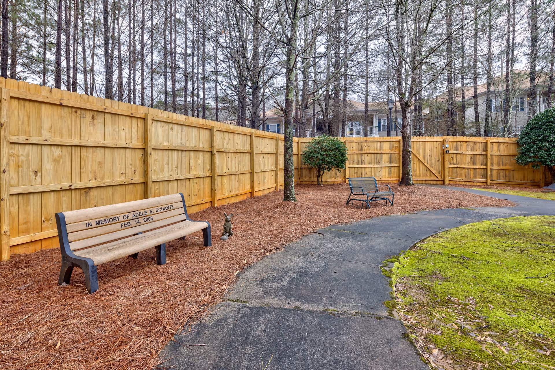 Outdoor area with a curved paved walkway, two wooden benches, a small statue of a rabbit, and a wooden fence surrounding the space. Trees and bushes are present along the fence, and residential buildings are visible in the background.