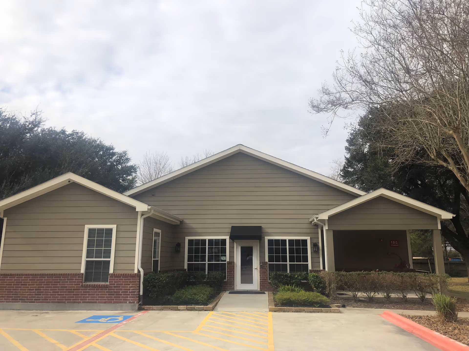 Exterior view of a single-story building with beige siding and a brick base, featuring a central entrance door with a small black awning above it, flanked by two large windows. There is a covered area on the right side and some bushes and trees surrounding the building. The sky is cloudy and there is a handicapped parking space in front.