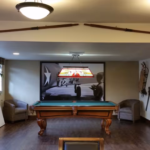 A room with a pool table centered under a hanging light fixture. On the back wall is a large black and white photograph of a vintage airplane and a person. Two armchairs are positioned on either side of the pool table. The floor is wooden, and the ceiling has a round light fixture and two wooden paddles mounted above the opening.