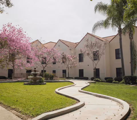 Outdoor courtyard area of a senior living facility with a winding concrete pathway, green grass, blooming pink trees, palm trees, and a multi-tiered stone fountain in front of a beige two-story building with a red-tiled roof.