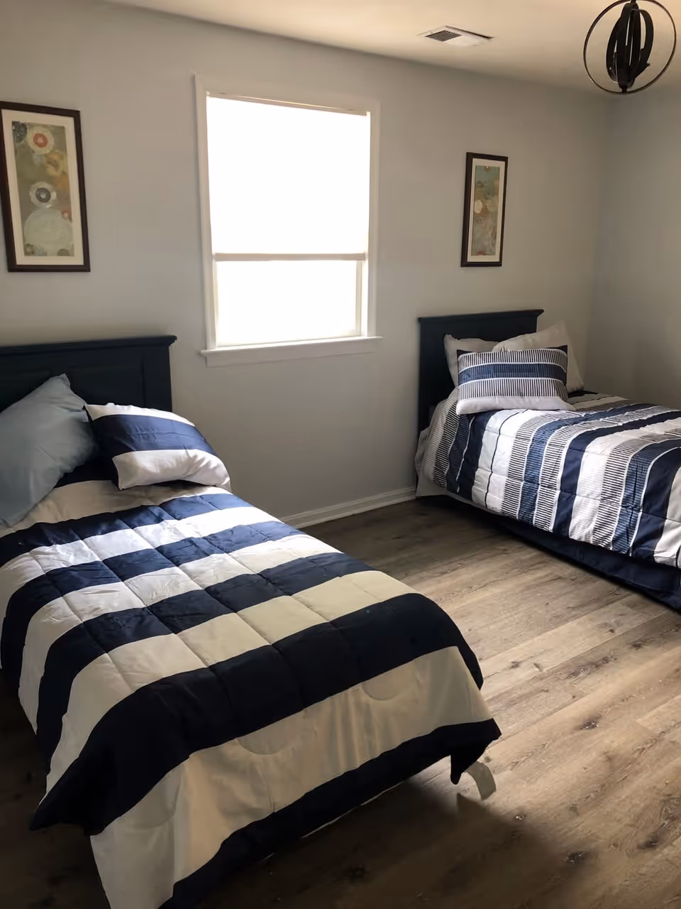 A bedroom with two twin beds, each with blue and white striped bedding and pillows. There is a window with a white blind between the beds, two framed abstract artworks on the wall, and a modern black ceiling light fixture. The floor is wood with a light finish.