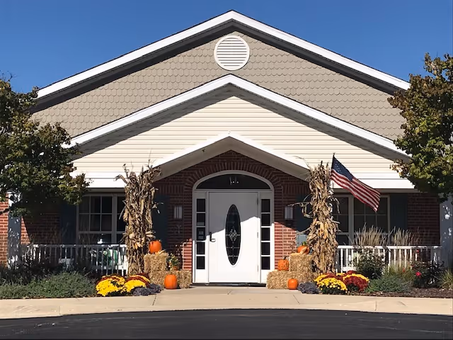 Front exterior view of a single-story building with a white door and a small porch decorated with hay bales, pumpkins, corn stalks, and colorful flowers. An American flag is displayed on the right side of the porch. Trees flank both sides of the building under a clear blue sky.