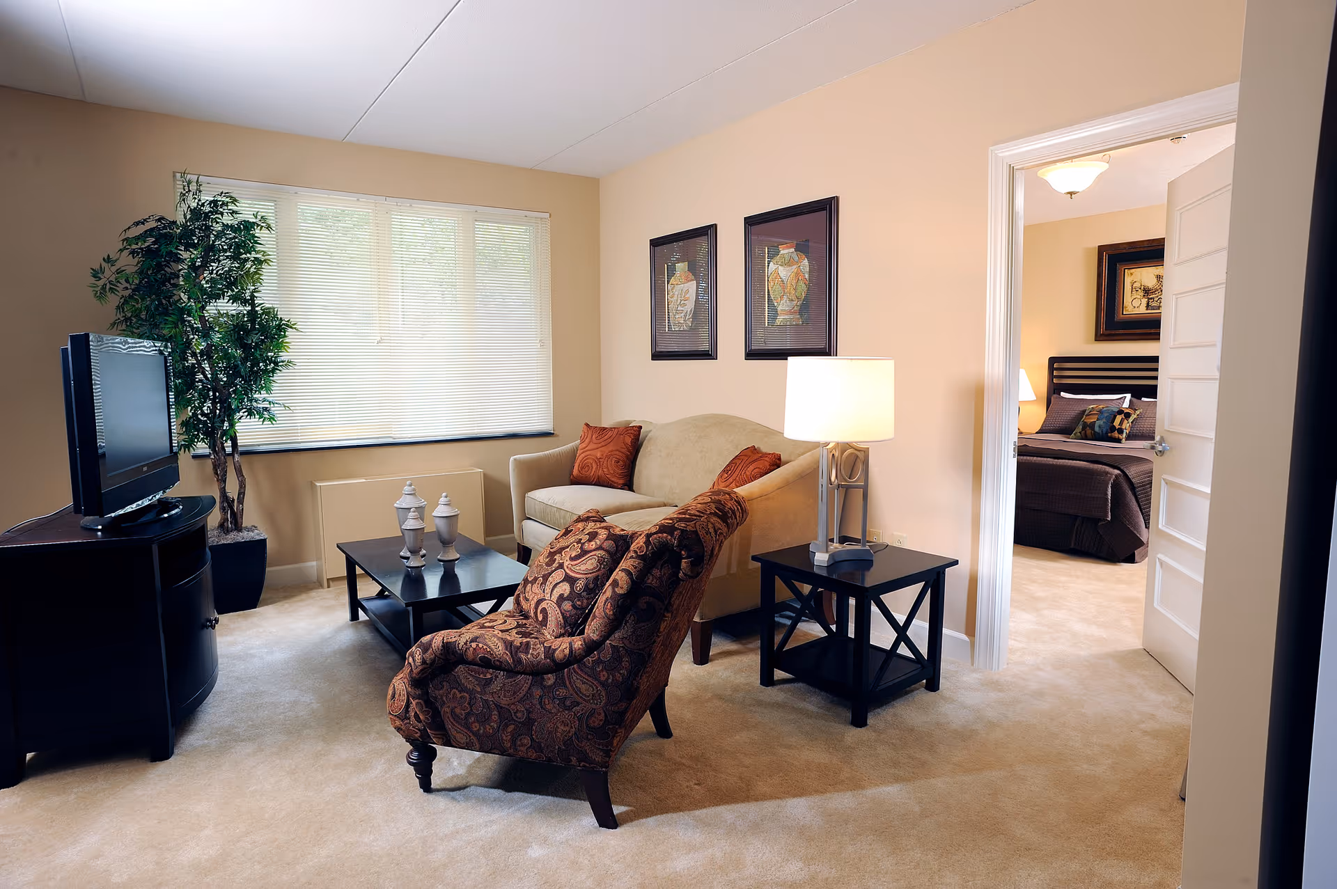 A cozy living room with a beige sofa, patterned armchair, black coffee table with decorative items, a side table with a lamp, a flat-screen TV on a black TV stand, and a large window with blinds. A doorway leads to a bedroom with a bed and framed artwork on the wall.