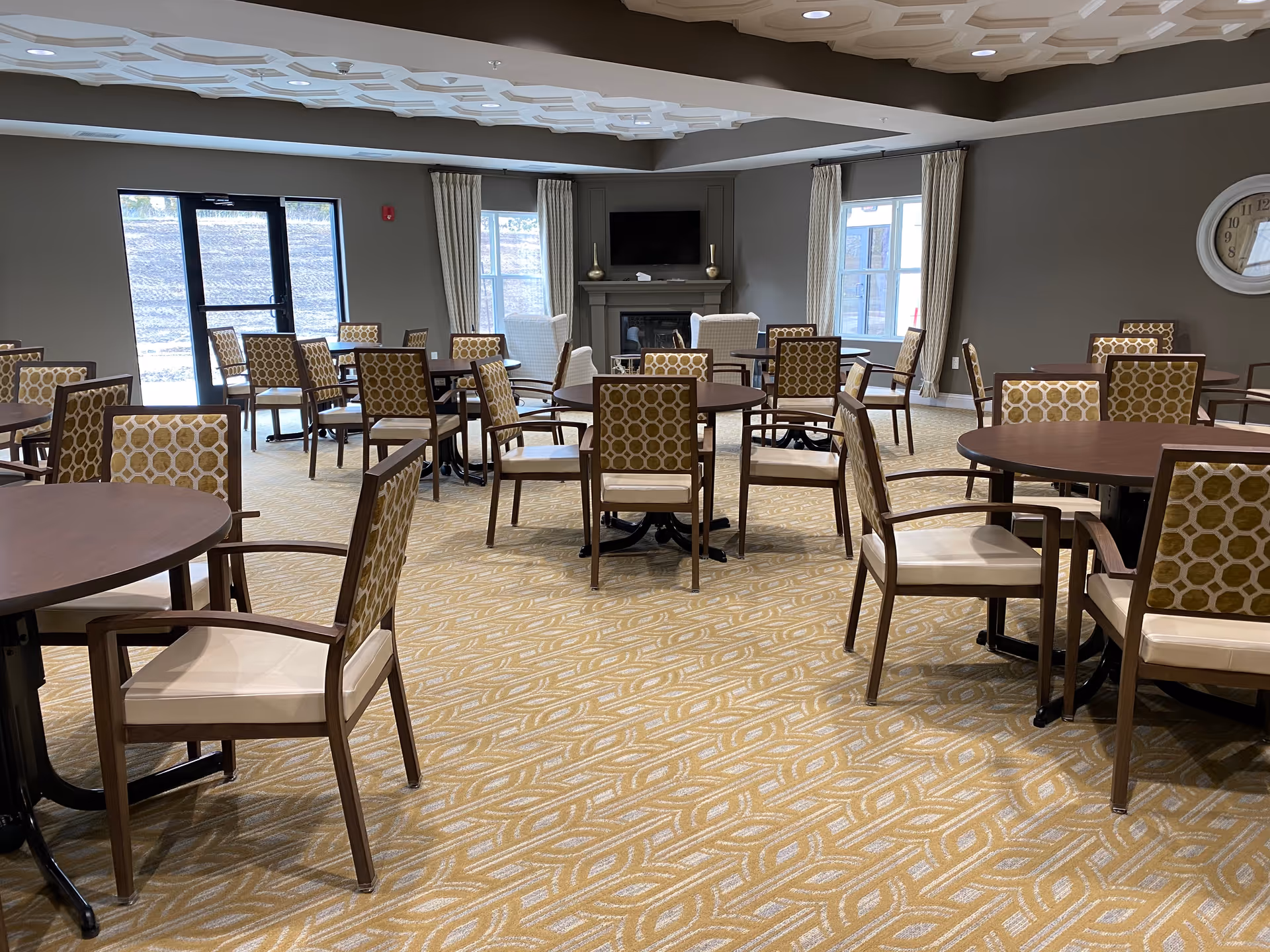 Spacious dining room with multiple round tables and patterned chairs facing a TV and fireplace.
