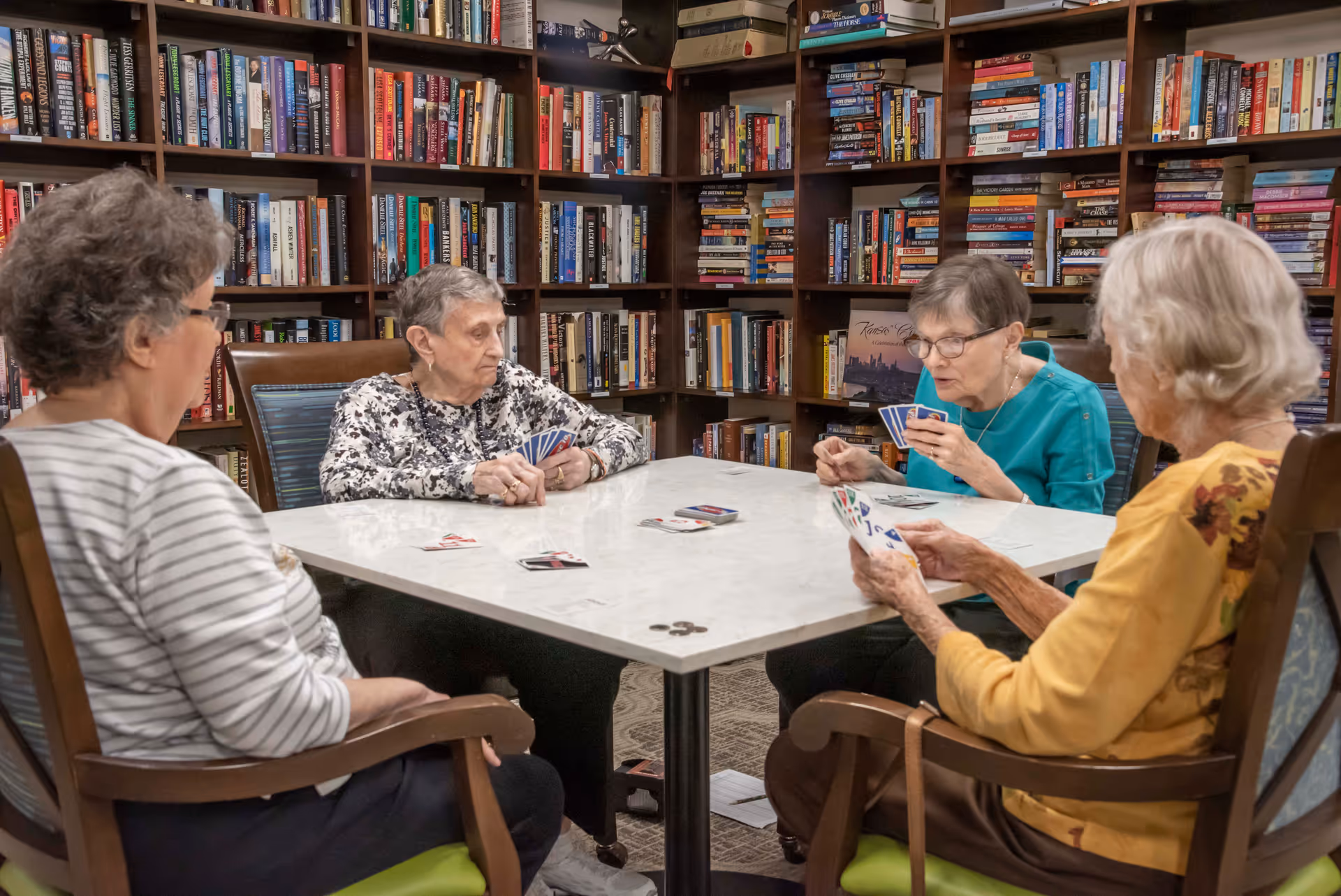 Four elderly women sitting around a square table playing cards in a room with bookshelves filled with books in the background.