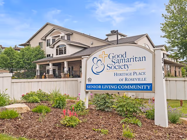 Exterior view of a senior living community building with a large sign in the foreground that reads 'Good Samaritan Society Heritage Place of Roseville Senior Living Community'. The building has multiple windows, a porch, and is surrounded by greenery and a white fence.