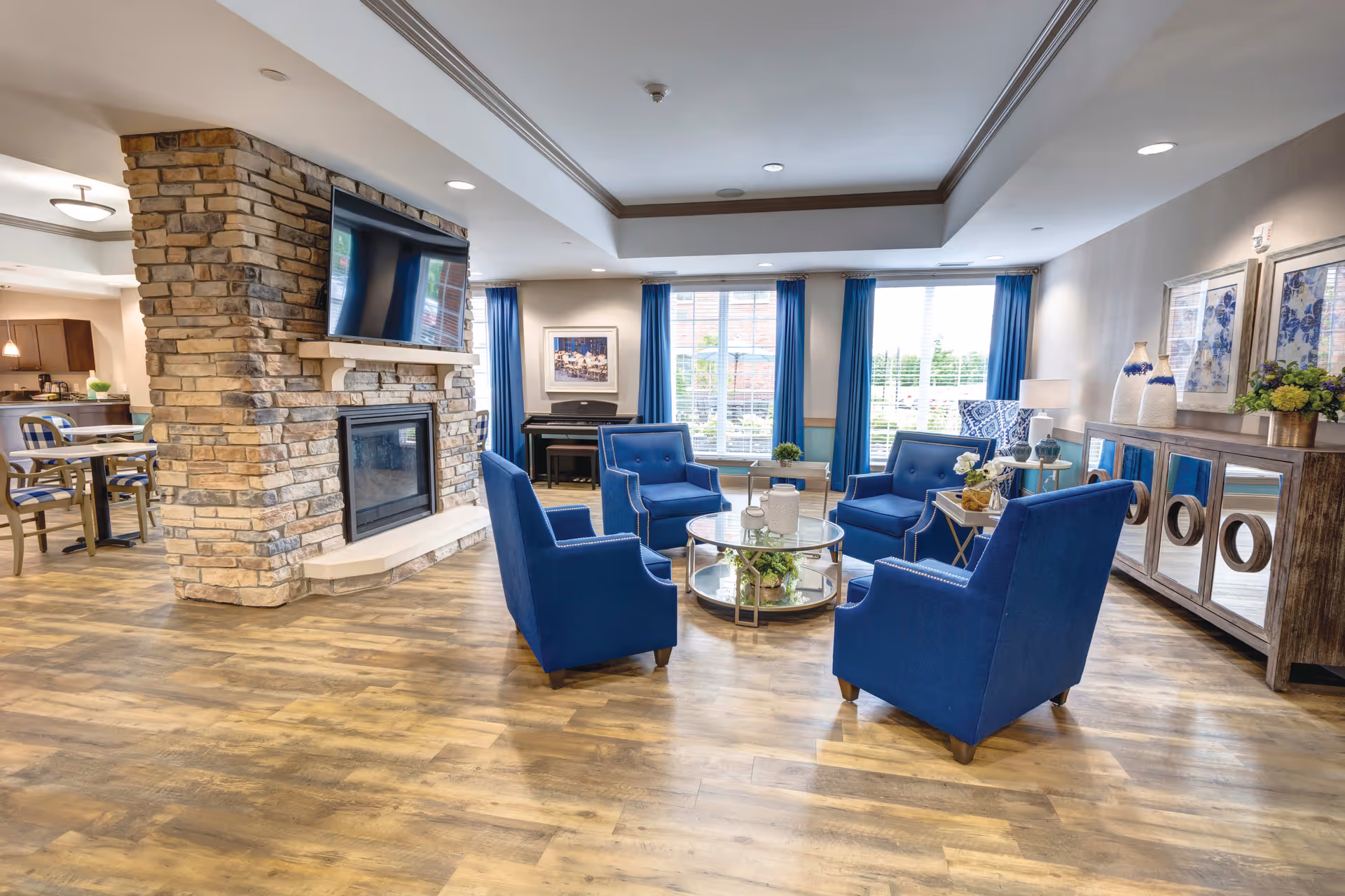 A bright and spacious senior living common area featuring a stone fireplace with a mounted flat-screen TV above it. Four blue upholstered armchairs are arranged around a round glass coffee table on a wood floor. Large windows with blue curtains let in natural light. A piano is visible in the background, along with a sideboard decorated with vases and framed artwork on the walls.